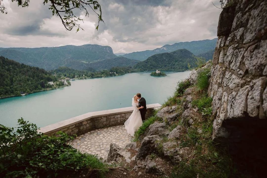 A bride and groom embracing on a stone path with a scenic view of Lake Bled, the island, and surrounding mountains in the background.