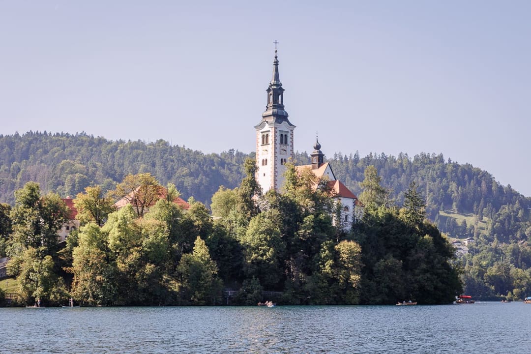 A view of the Church of the Assumption on an island in Lake Bled, surrounded by lush greenery and calm water.