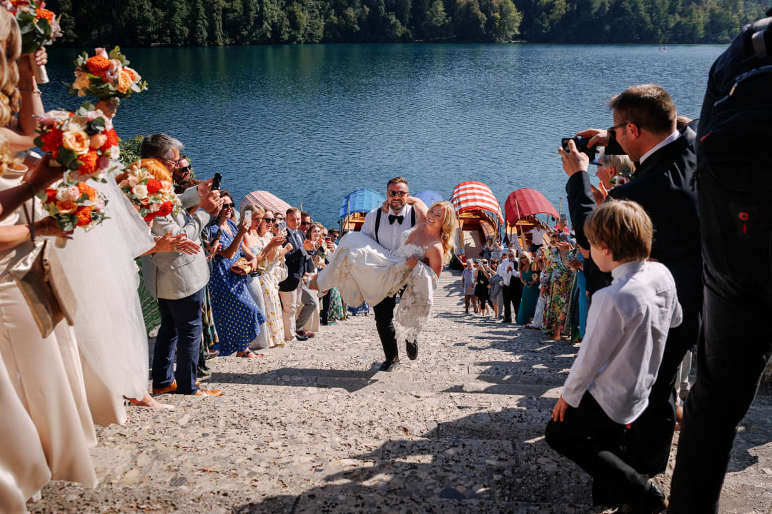 A groom carrying the bride up steps surrounded by cheering guests, with Lake Bled and colorful boats in the background.
