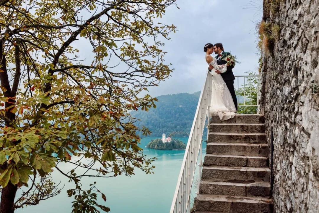 A bride and groom sharing a kiss on stone stairs with Lake Bled and its island church visible in the background.