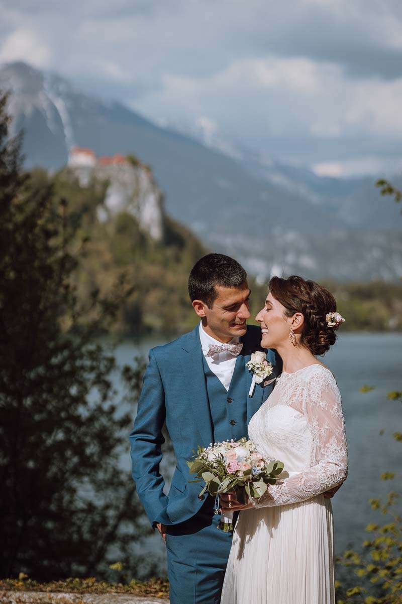 A bride and groom smiling at each other with Lake Bled and a castle in the background.