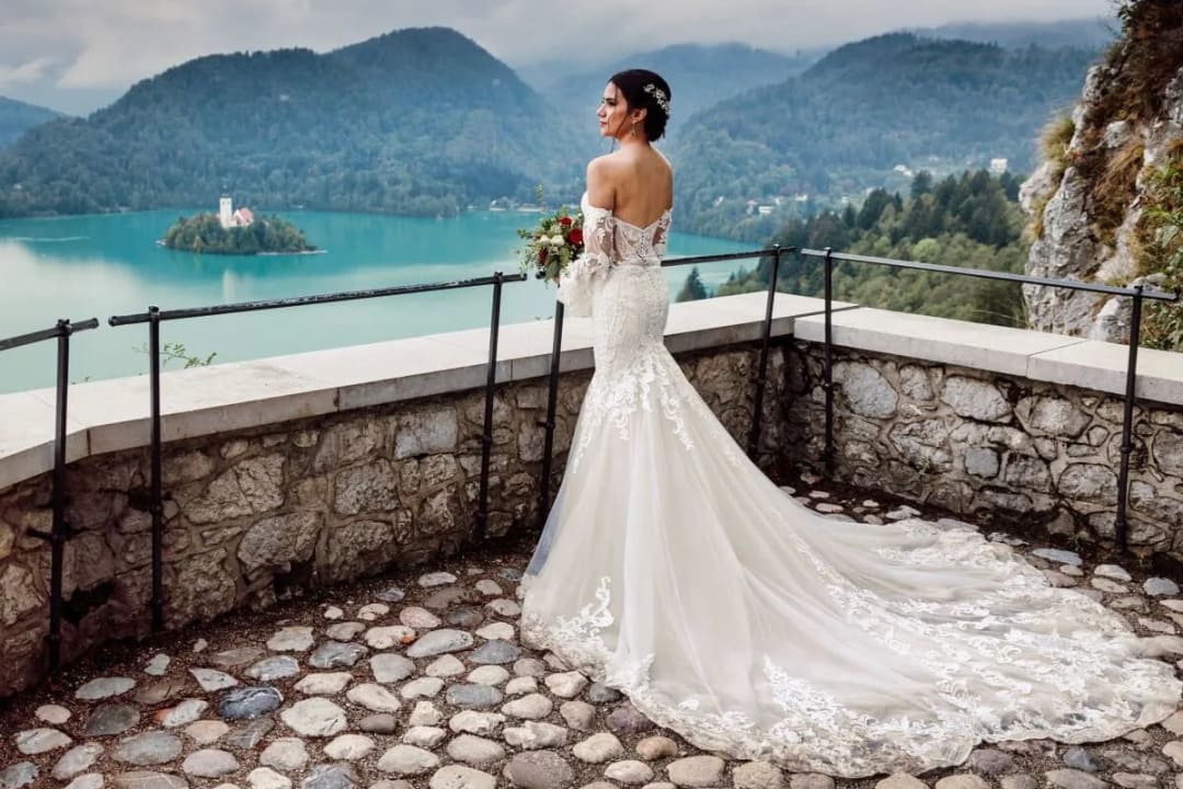 A bride in a lace wedding dress stands on a stone terrace, overlooking the scenic Lake Bled and its island.