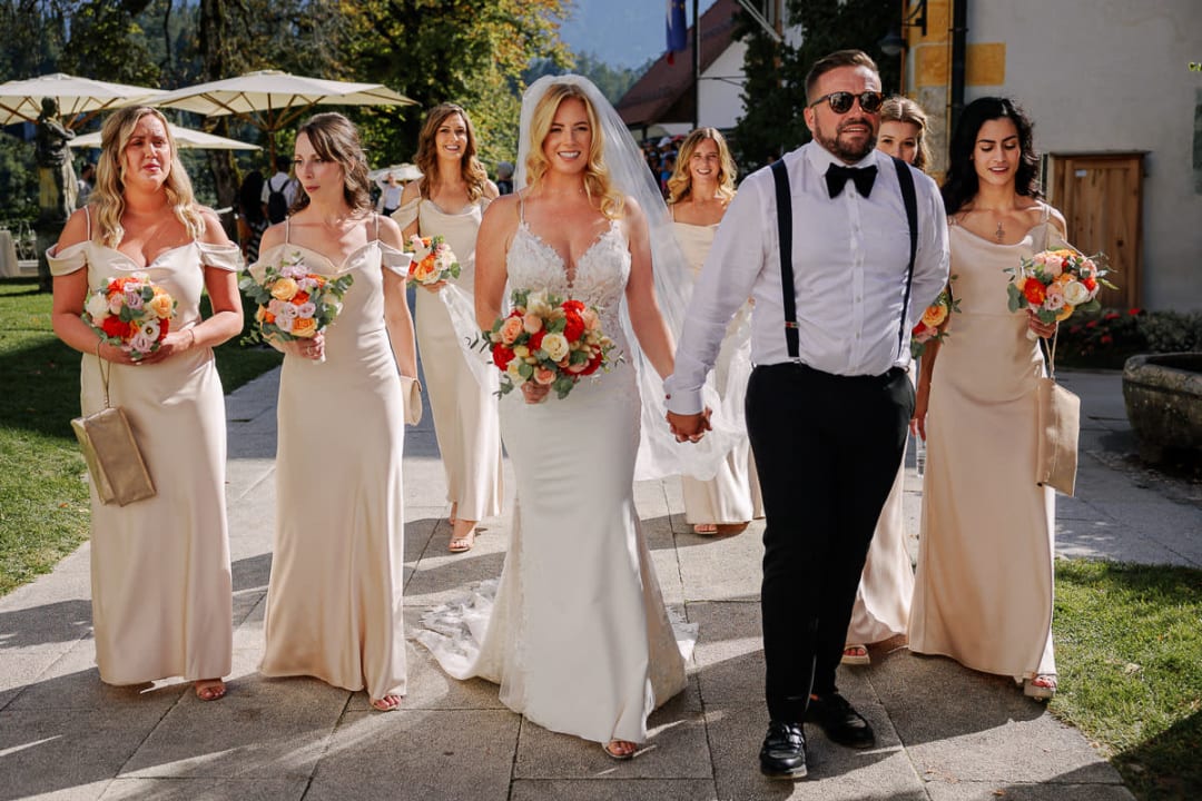 A bride and groom walking hand in hand with bridesmaids in matching dresses carrying bouquets on a sunny day.