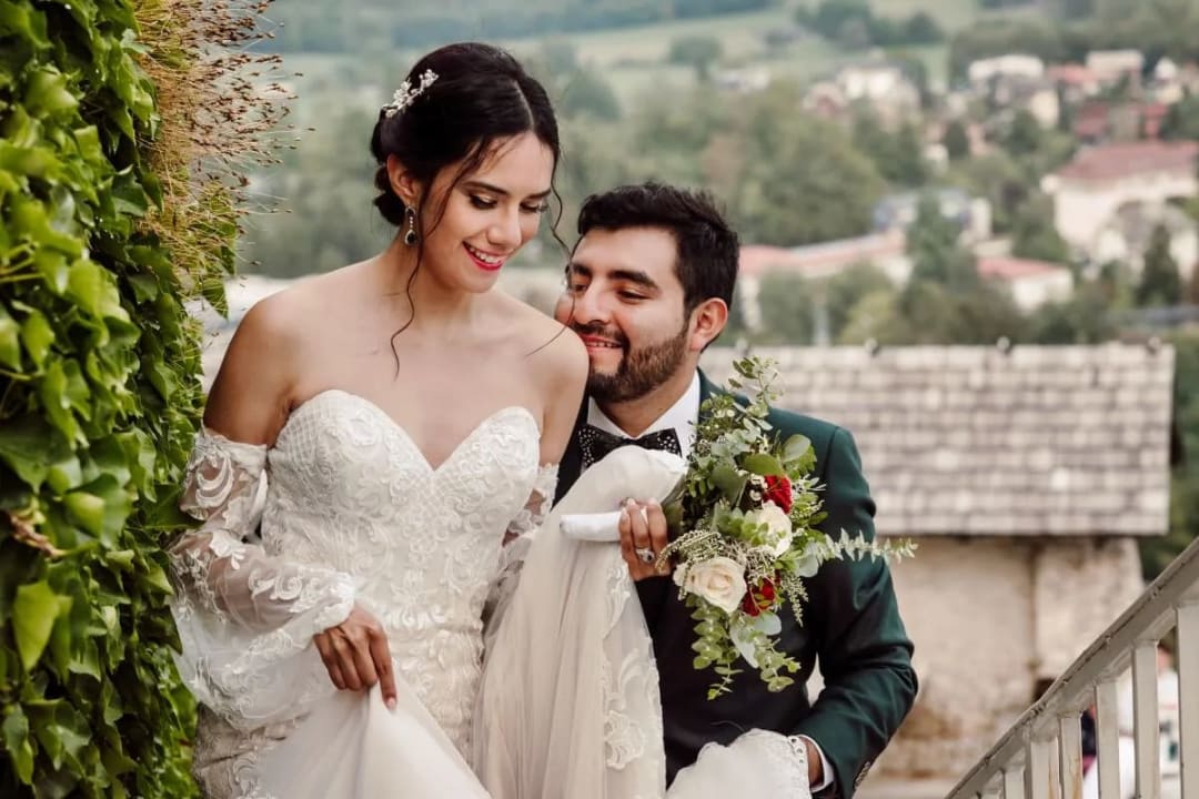 A groom lovingly supporting the bride as they ascend a staircase, surrounded by greenery, with a scenic view in the background.