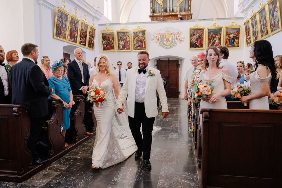 A bride and groom smiling as they walk down the aisle of a church, surrounded by family and friends.