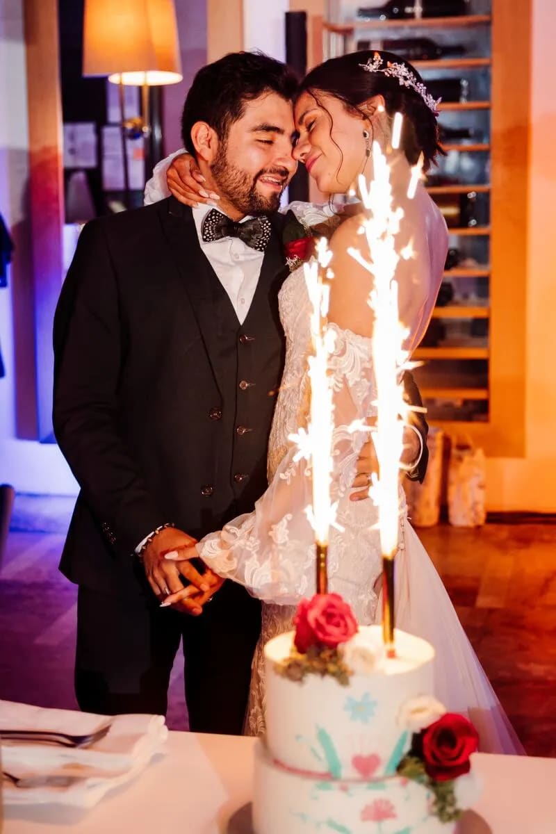 A bride and groom smiling and embracing in front of a wedding cake with sparklers lit on top.