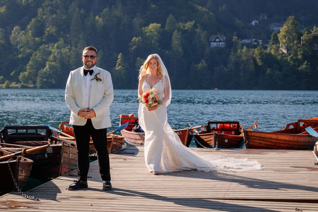 A bride and groom standing on a dock with wooden boats and a scenic lake view in the background at Lake Bled.