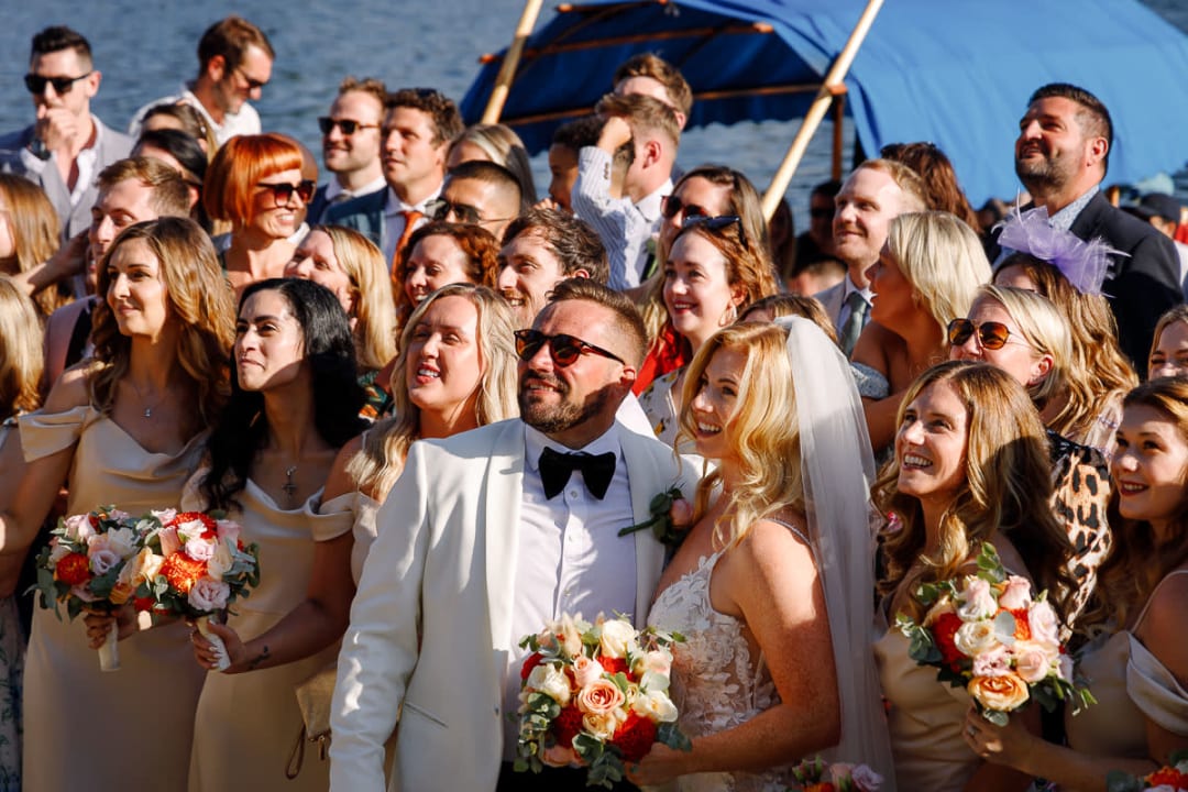 A bride and groom surrounded by smiling wedding guests posing together near a lake.
