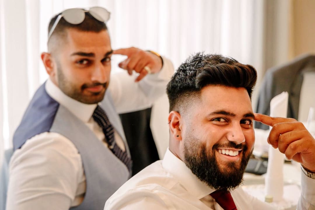 Two wedding guests in formal attire sharing a smile and gesturing towards each other indoors.