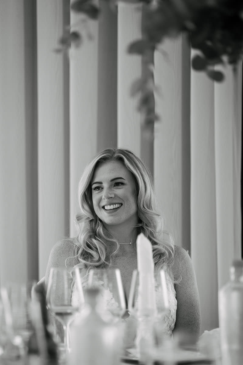A smiling bride sitting at a table during a wedding reception, surrounded by glasses and candles.