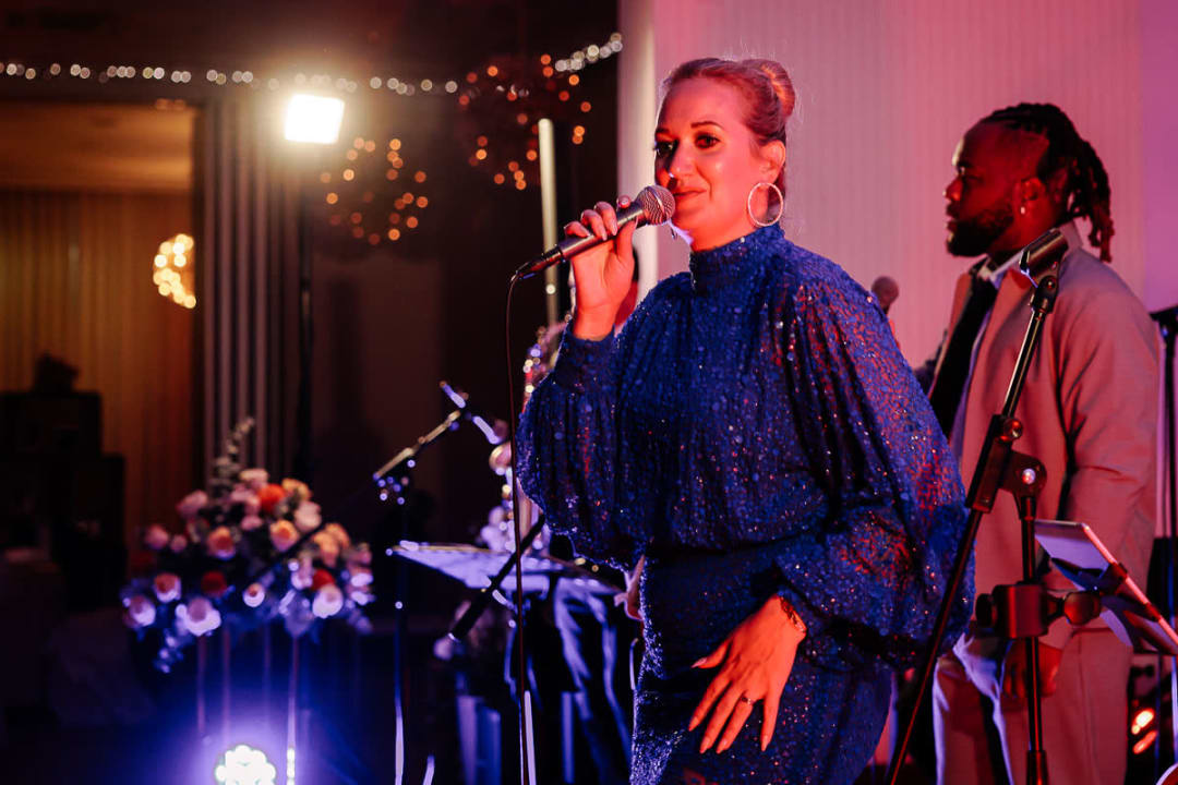 A singer in a blue dress performs at a wedding reception, with a microphone in hand and a musician in the background.