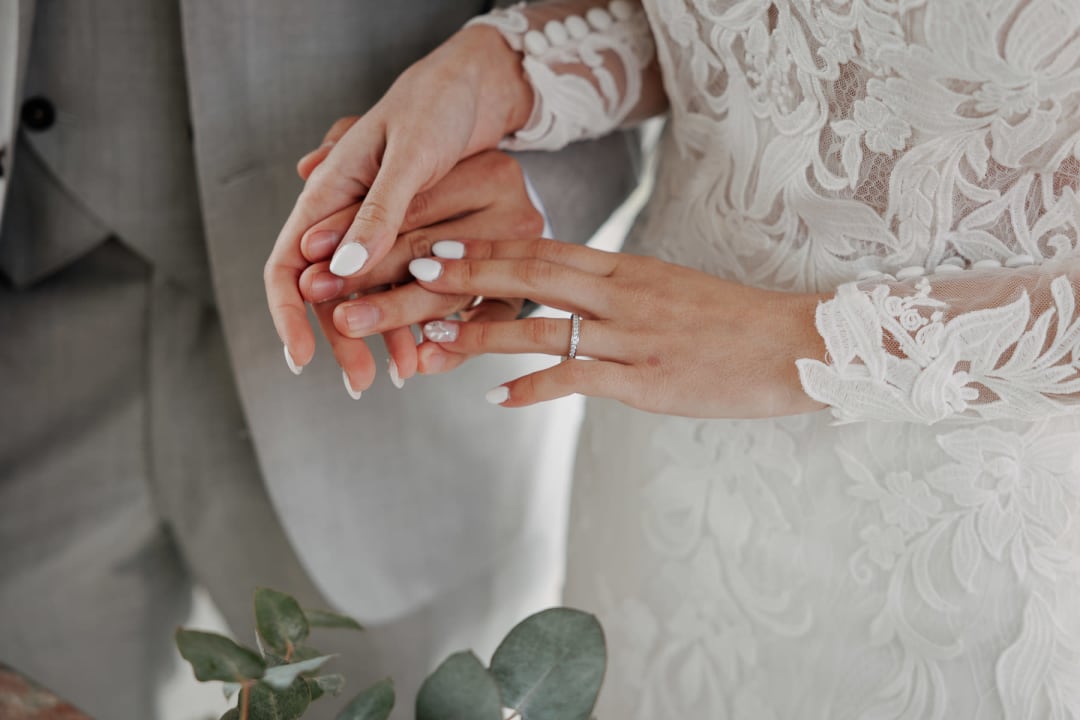 Close-up of hands during the ring exchange, showing the delicate lace sleeve of the bridal gown.