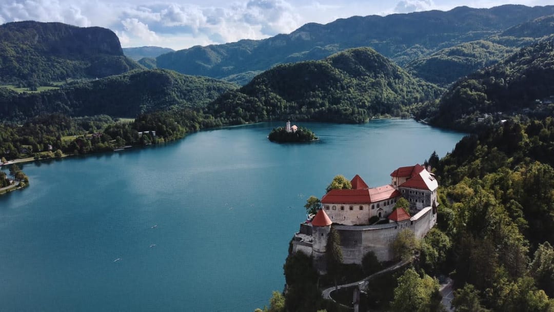 A scenic view of Lake Bled featuring a historic castle on the hill in the foreground and an island with a church in the middle of the lake, surrounded by lush mountains.