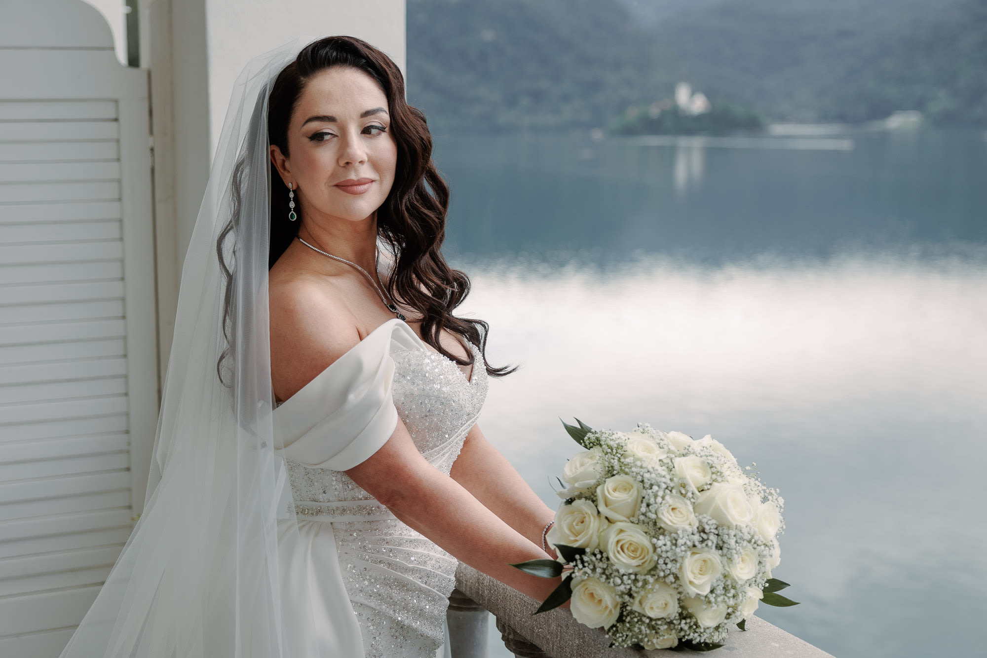 Bride on a balcony overlooking Lake Bled, elegant portrait with bouquet.