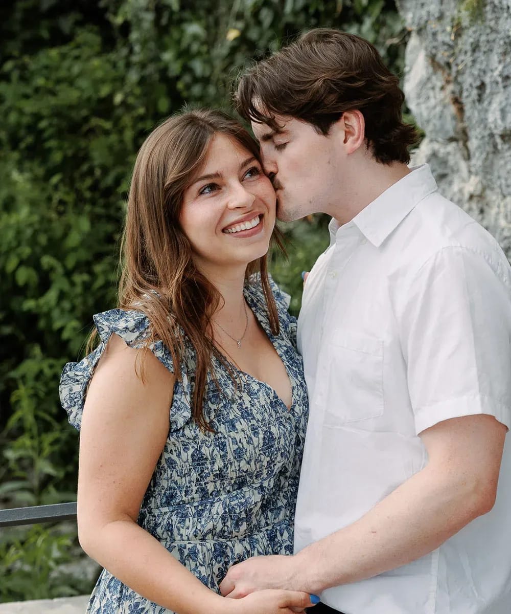 A smiling woman in a blue floral dress receives a gentle kiss on the cheek from her partner in a white shirt, holding hands in a romantic outdoor setting.