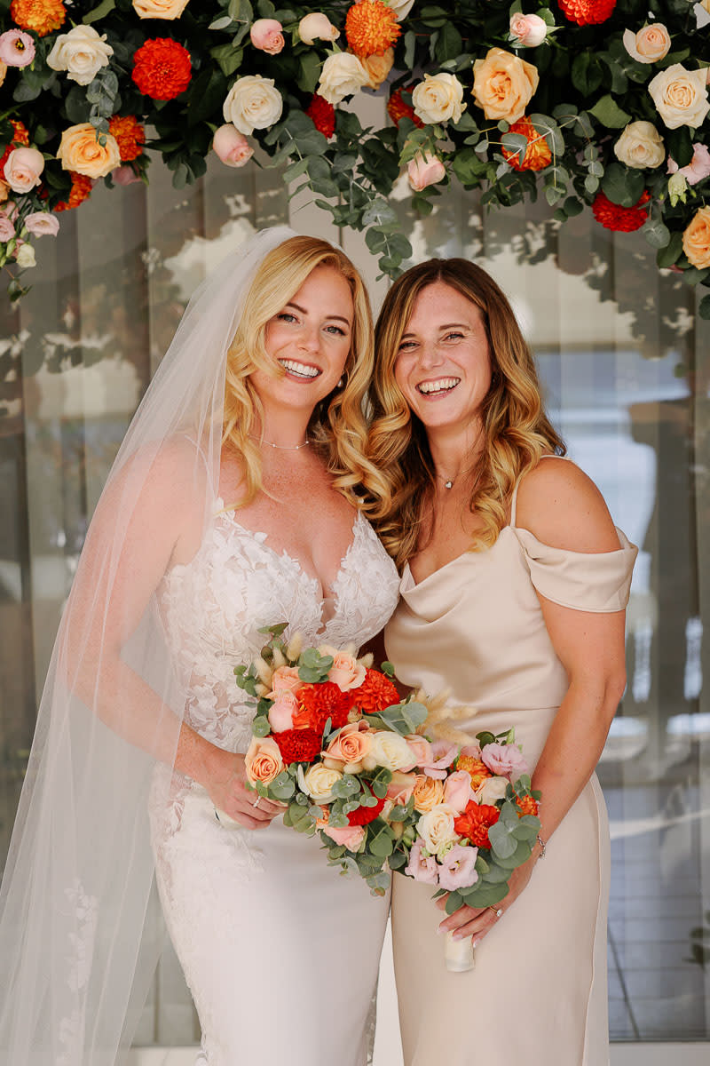 A smiling bride in a lace gown stands with a bridesmaid under a vibrant floral arch, each holding colorful bouquets.