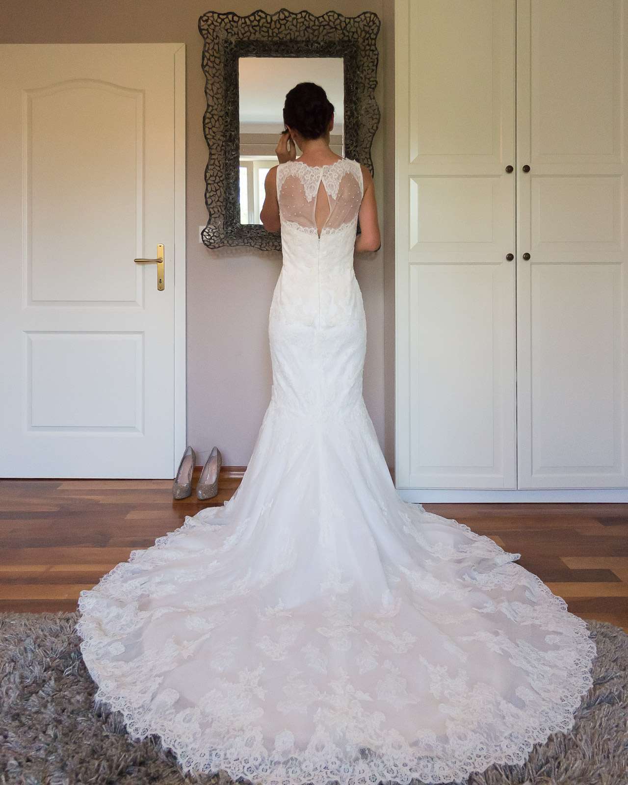 A bride in a white lace wedding gown standing in front of a mirror in a dressing room, adjusting her makeup.