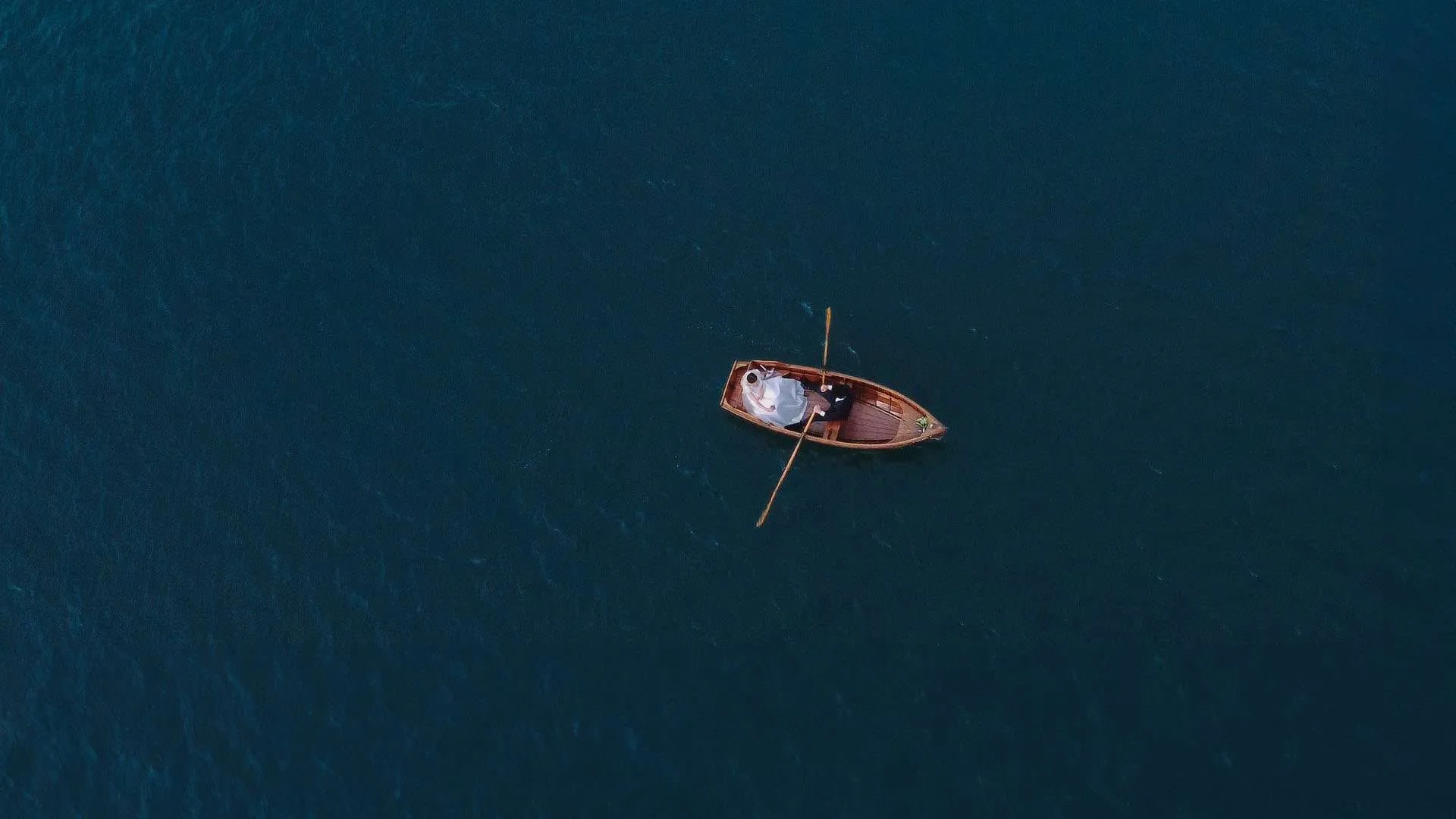 Aerial view of a bride and groom in a wooden boat on a serene lake.
