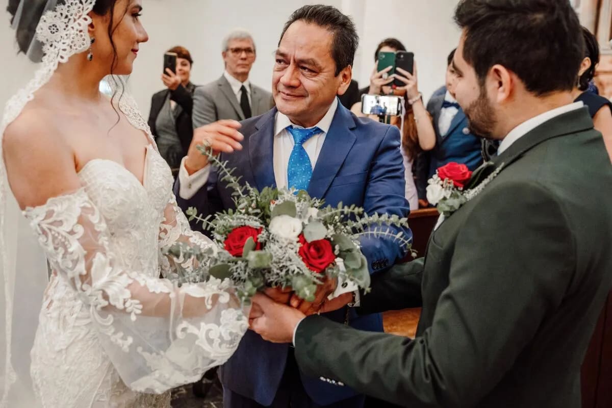 A touching moment as a man in a blue suit hands a bouquet to the bride and groom during a wedding ceremony, surrounded by guests capturing the scene.