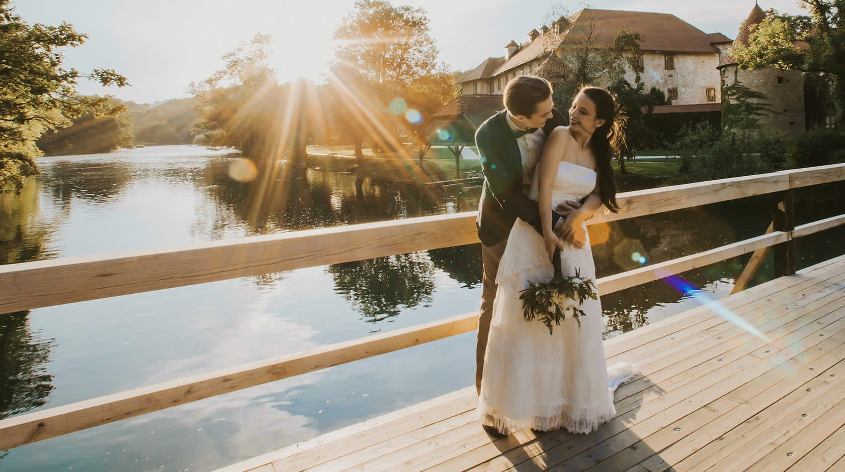 A bride and groom share a joyful moment on a wooden bridge as the sun sets, casting a warm glow over a tranquil lake and a historic building in the background.