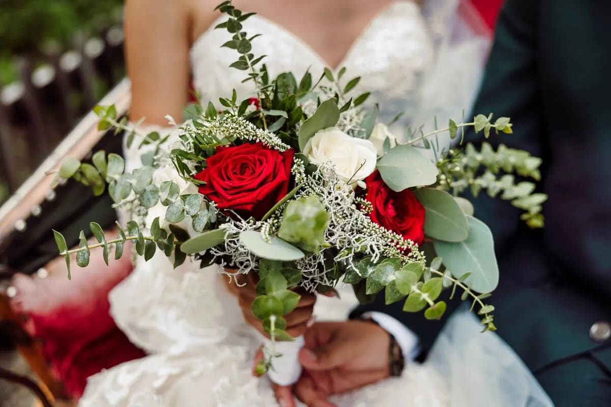 A bride and groom seated together, holding a beautiful bouquet of red and white roses with greenery.