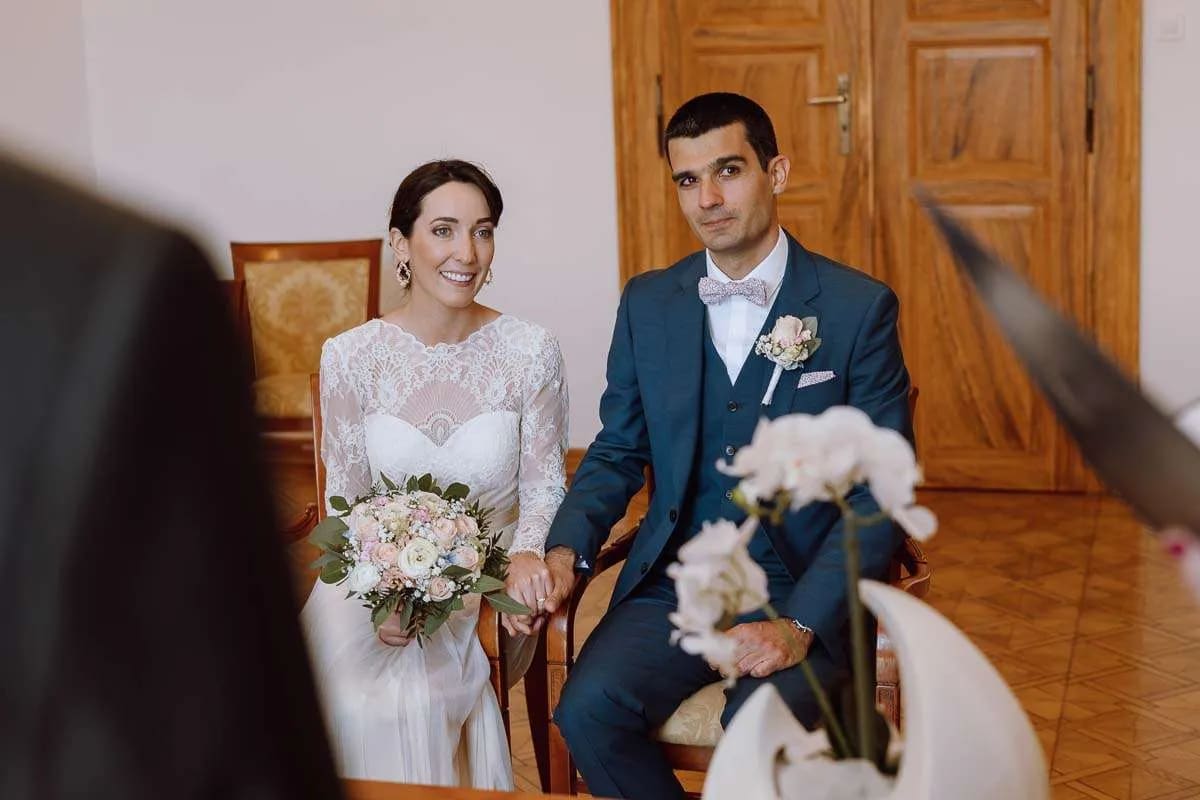 A bride and groom sit together during a wedding ceremony, holding hands and smiling, with a bouquet in the bride's lap.