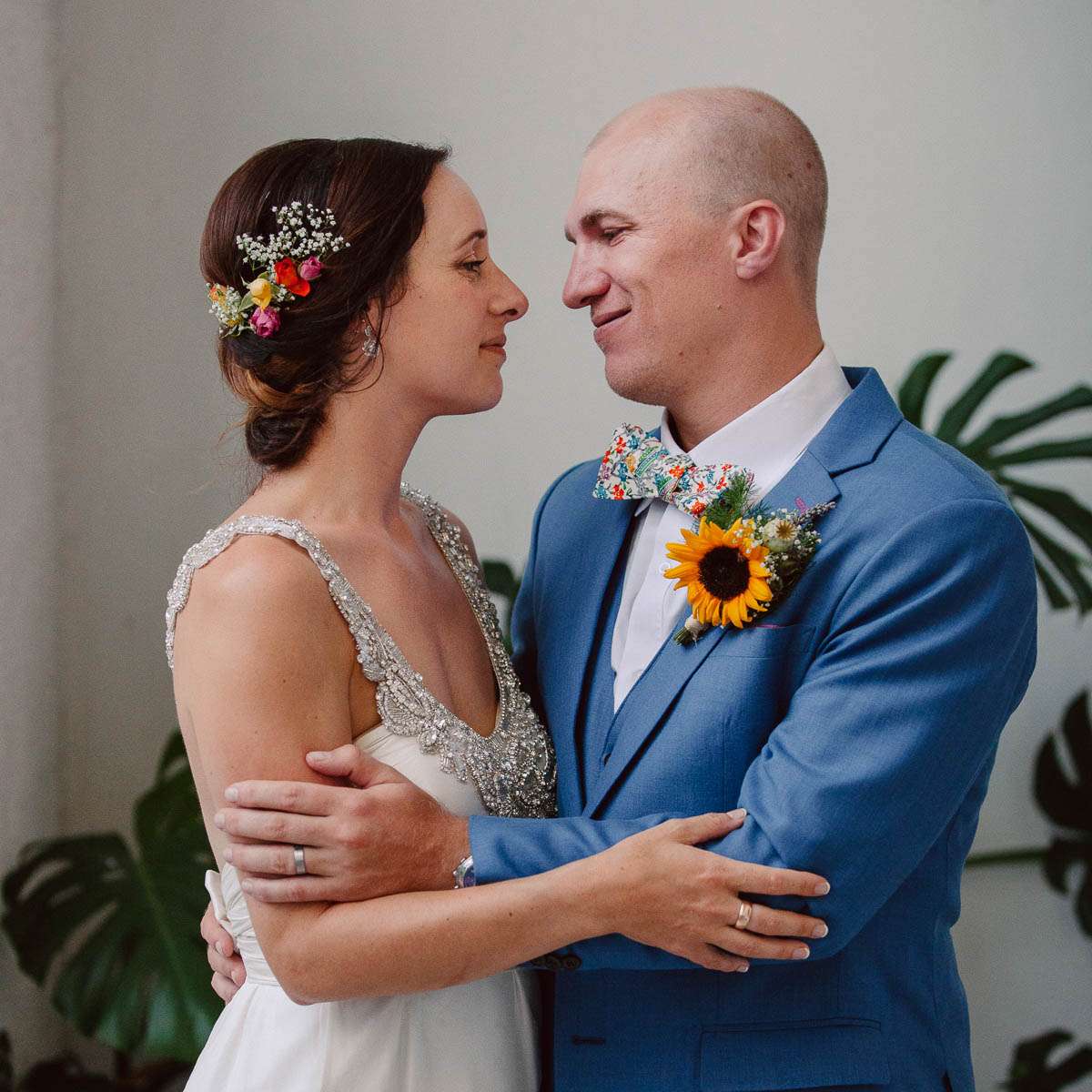 A bride and groom lovingly facing each other, embracing indoors. The bride wears an elegant dress adorned with intricate beading, and the groom wears a blue suit with a sunflower boutonniere.
