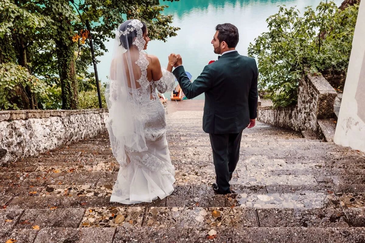 A bride and groom walk hand in hand down stone steps surrounded by greenery, with a view of a serene lake in the background.