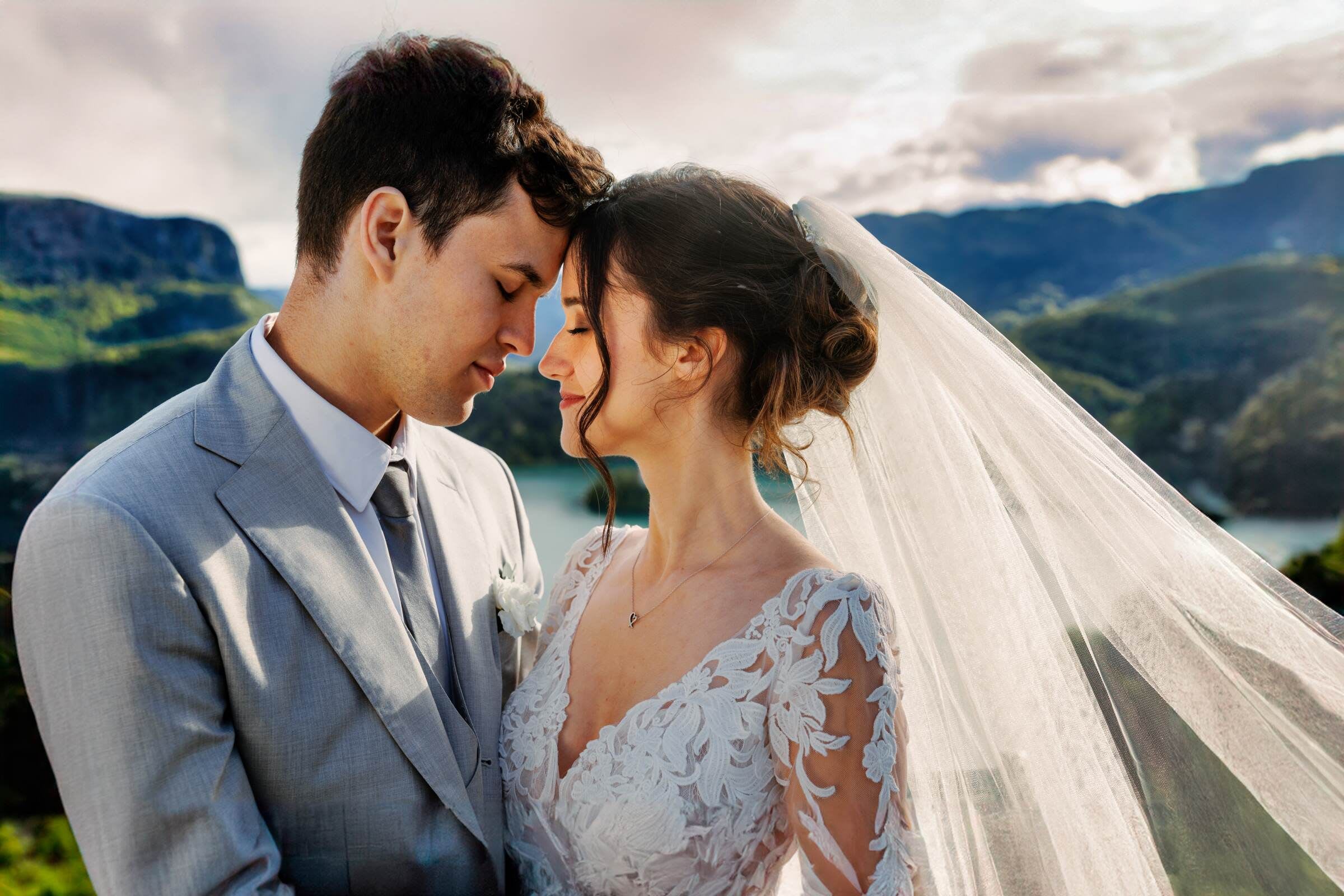 A bride and groom standing close together, eyes closed, with a scenic mountain and lake view in the background.