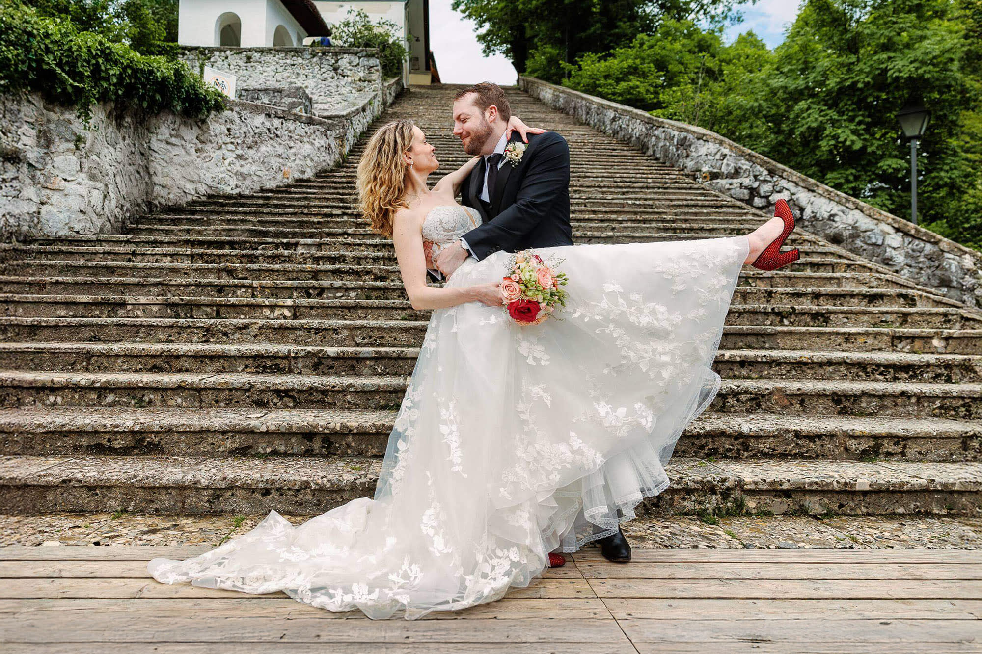 A groom holds the bride in his arms as they pose joyfully on stone steps. The bride is holding a bouquet of flowers and wearing red shoes.