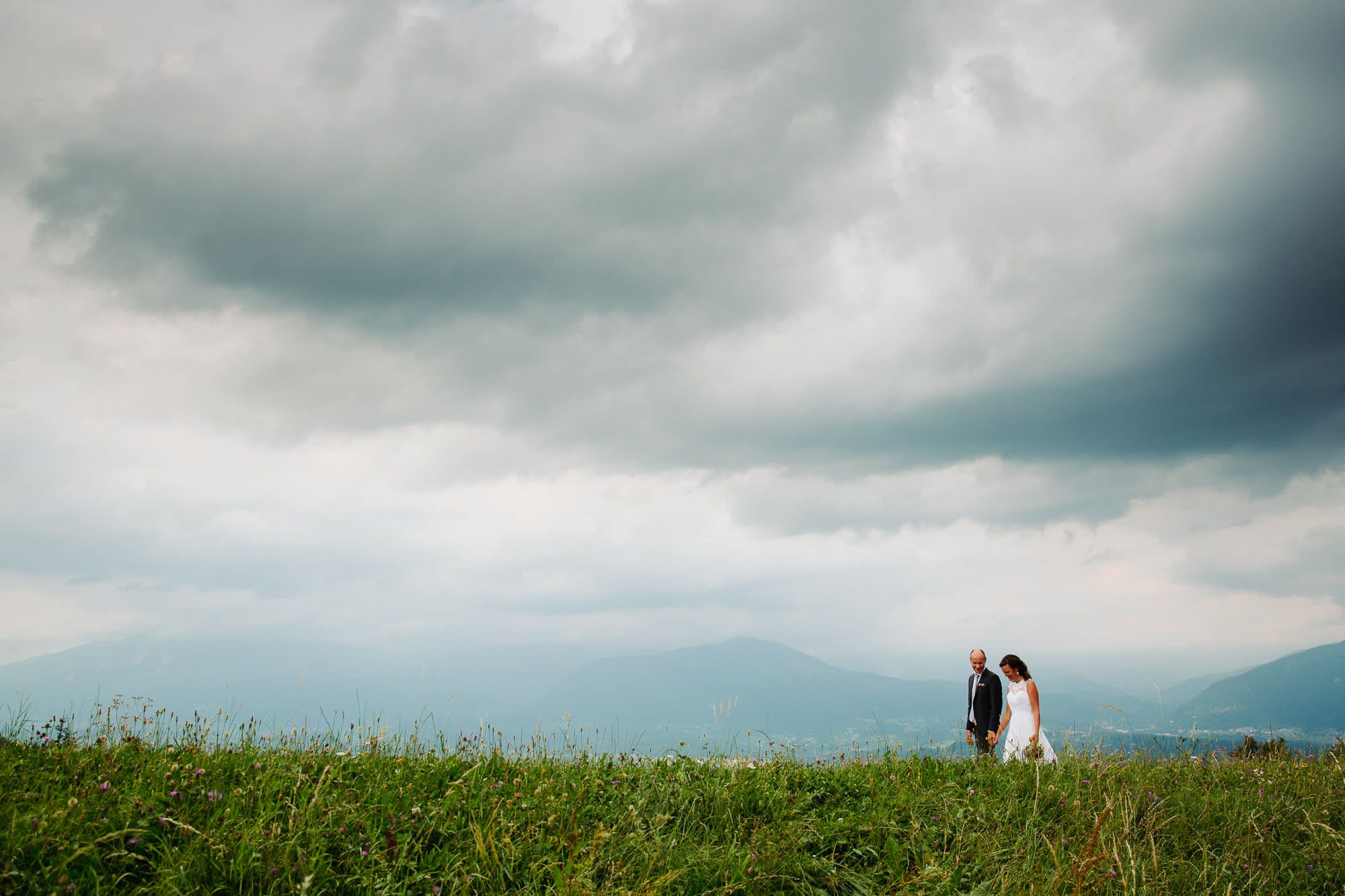 A bride and groom walking hand in hand in a field with dramatic clouds and mountains in the background.