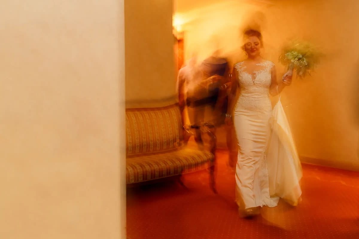 A bride in a white lace gown holding a bouquet, walking indoors with a soft, blurred effect.