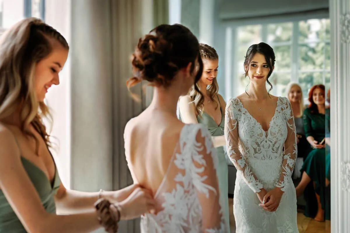 A bride smiling while a bridesmaid helps her with the wedding dress, surrounded by friends in a bright room.