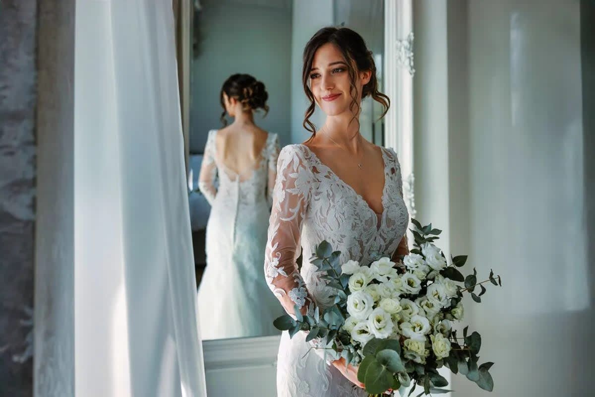 A bride in an elegant lace wedding dress stands smiling while holding a bouquet of white flowers, with her reflection visible in a mirror.