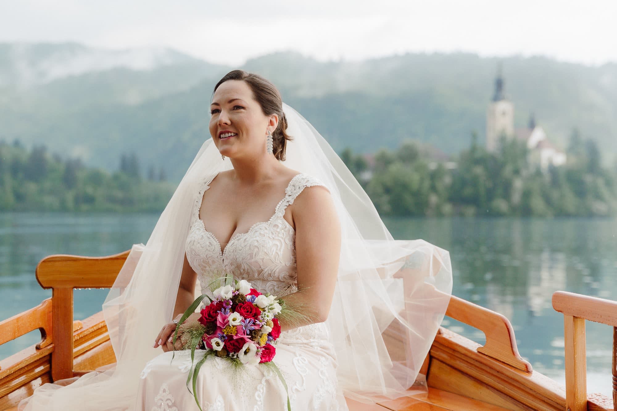 A bride sitting in a wooden pletna boat on Lake Bled with her veil, bouquet, and the island church softly visible in the background.