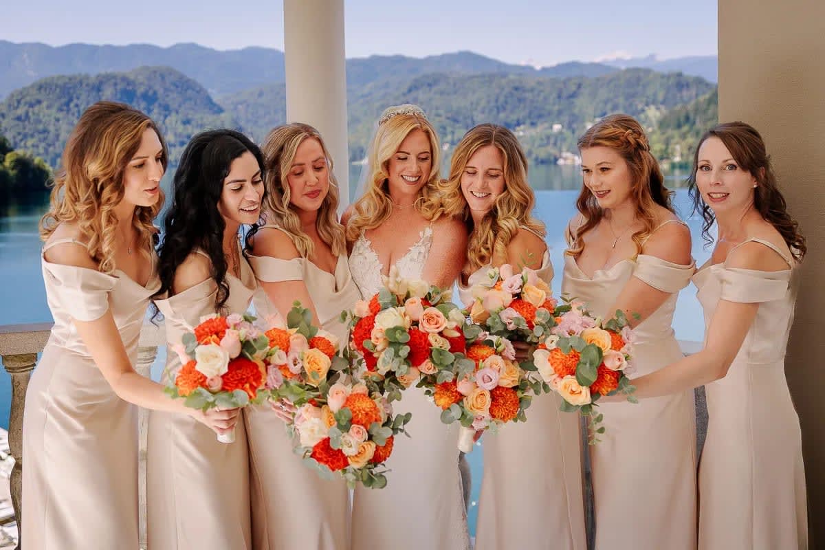 A bride and her bridesmaids in elegant dresses holding vibrant bouquets, with a view of Lake Bled and mountains in the background.