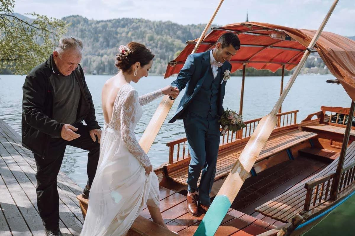 A bride and groom boarding a traditional wooden boat on a lake, assisted by an older man.