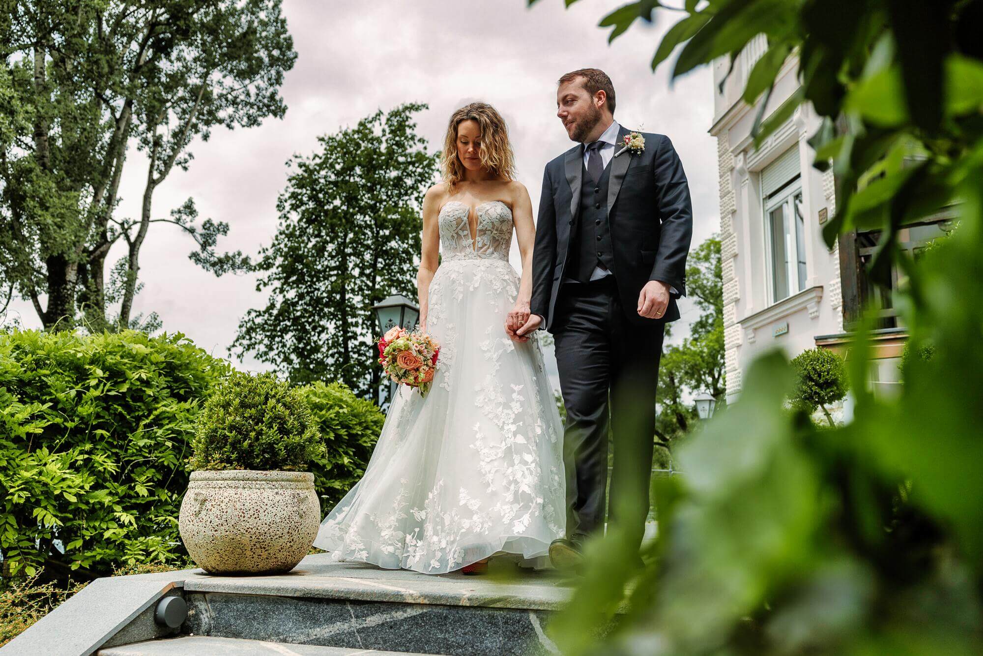 A bride and groom holding hands and walking down steps surrounded by lush greenery and flowers.