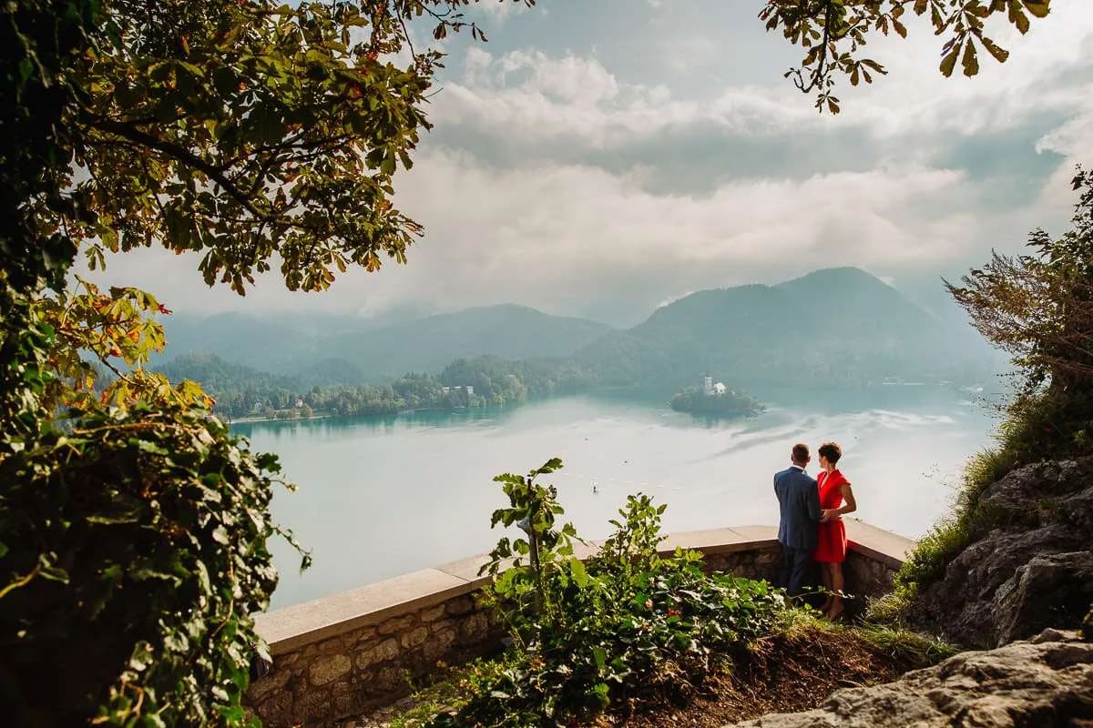A couple stands together, overlooking the scenic view of Lake Bled with mountains and an island in the background.