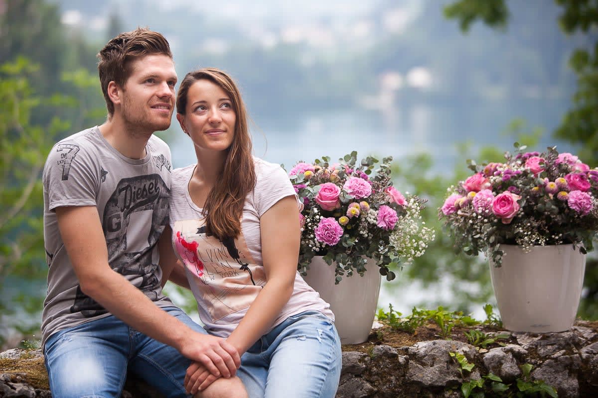 A couple sitting closely together, surrounded by colorful flower arrangements with a blurred view of Lake Bled in the background.