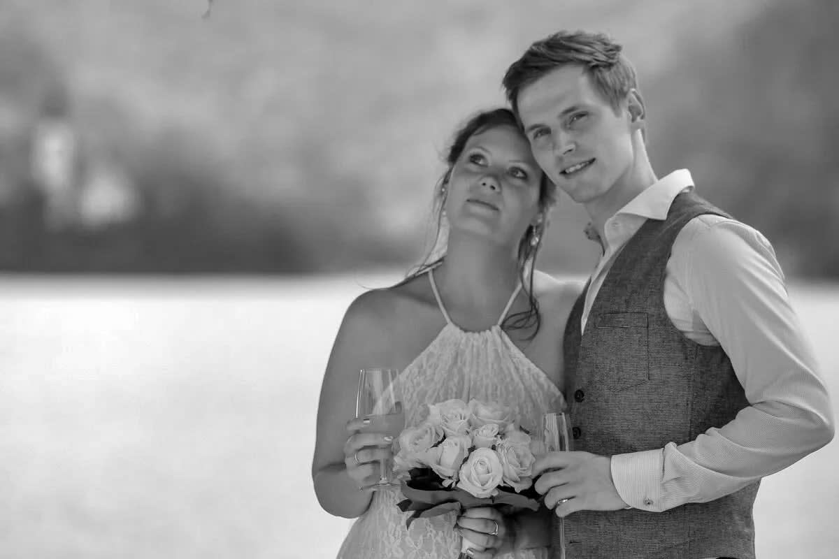 A bride and groom smiling and holding champagne glasses, with a blurred view of Lake Bled in the background.