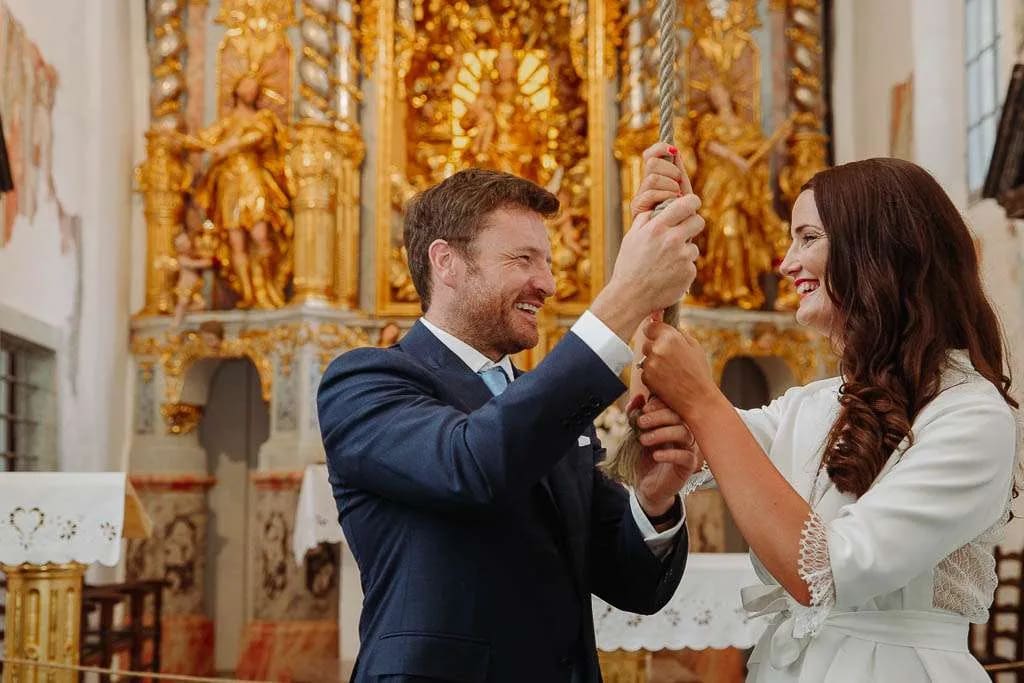 A bride and groom happily ringing the wishing bell together inside a beautifully decorated Bled island church.