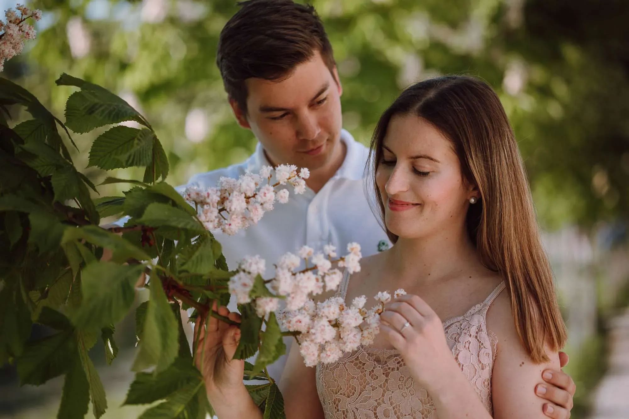 A young couple sharing a tender moment while admiring pink blossoms on a tree branch, with the man gently holding the woman from behind.