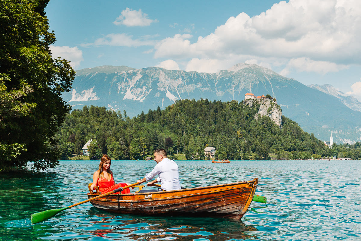 A couple enjoying a romantic ride in a rowboat on Lake Bled, with a backdrop of mountains and a distant view of Bled castle.