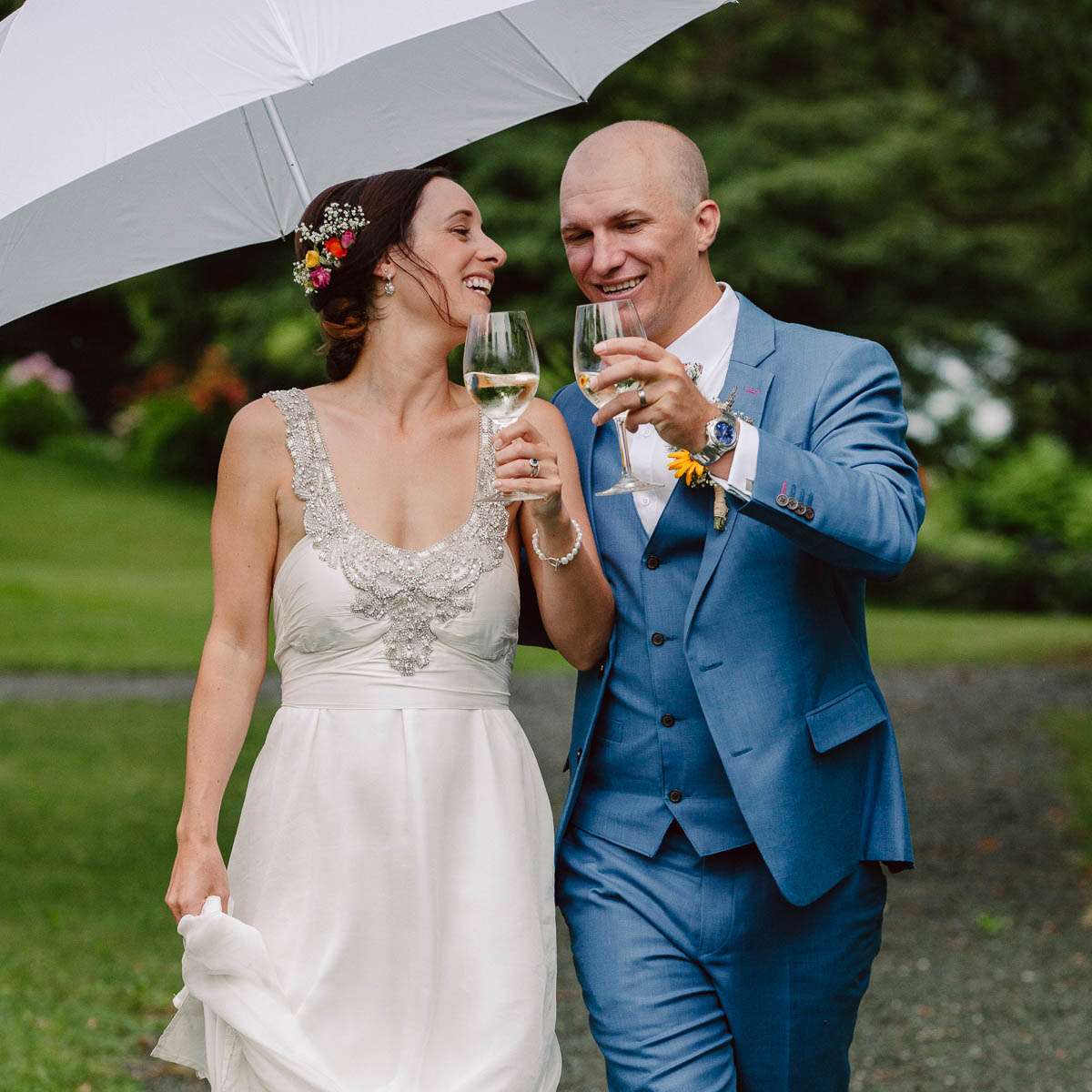 A bride and groom walking under an umbrella, toasting with wine glasses and smiling in a garden setting.