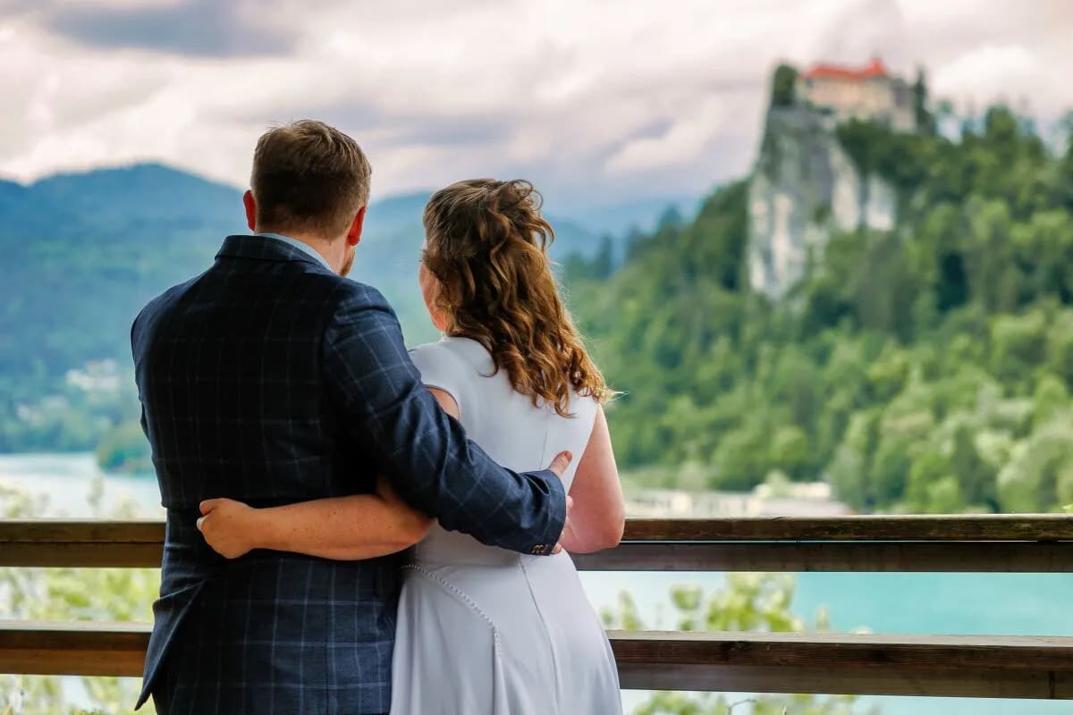 A couple standing together on a balcony, looking out at the scenic view of Lake Bled with a castle visible on a hill in the background.