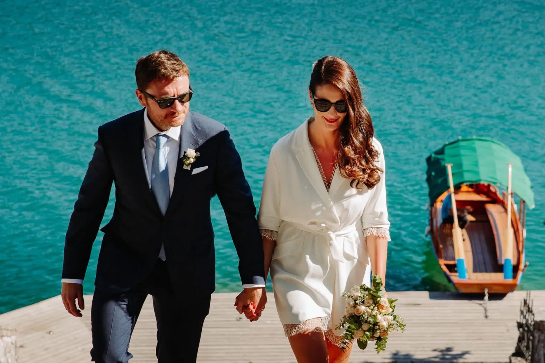 A bride and groom holding hands and walking up a dock with a wooden boat and the turquoise waters of Lake Bled in the background.