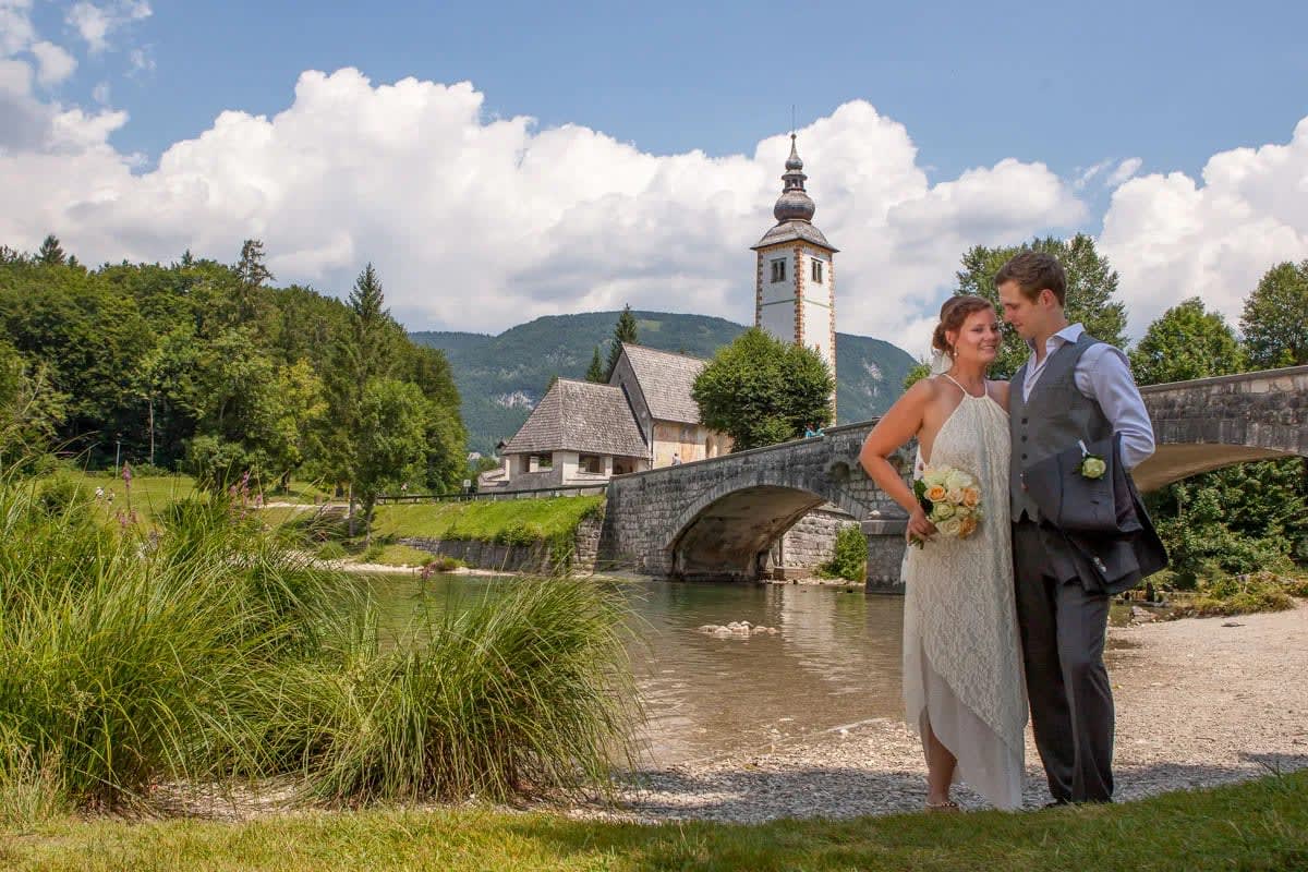 A bride and groom stand smiling by a stone bridge near a church, with mountains and trees in the background.