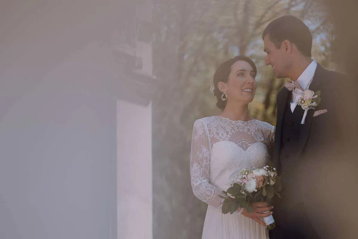 A bride and groom smiling at each other, with the bride holding a bouquet and wearing a lace dress.