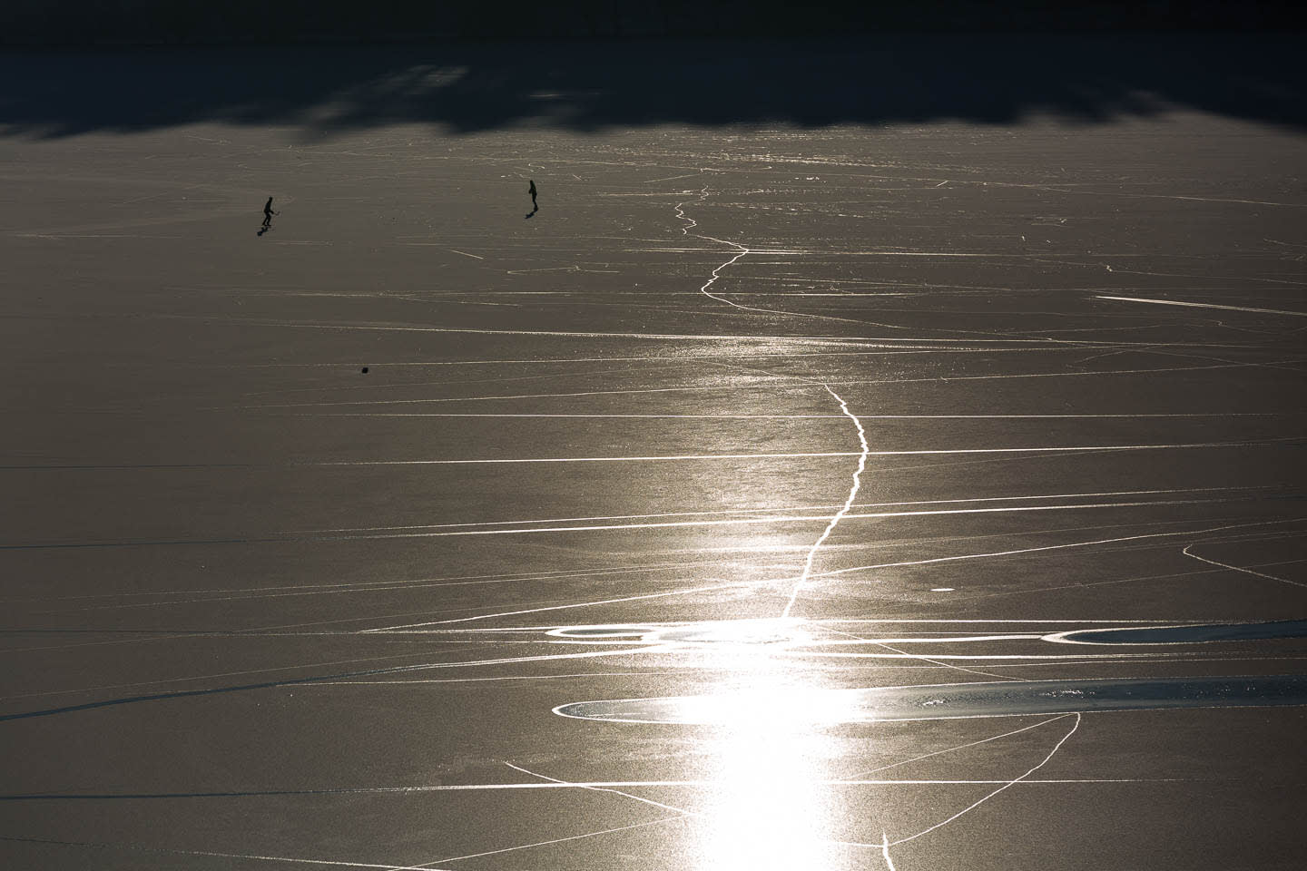 Two silhouetted figures ice skating on a frozen lake, with sunlight reflecting off the surface.